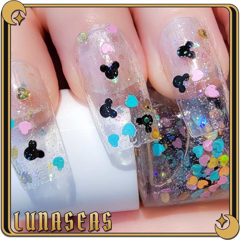 K&ouml;nnte beinhalten: Transparenter Nagellack mit holografischem Glitzer, kleinen pastellfarbenen herzf&ouml;rmigen Pailletten und schwarzen Akzenten in Form von Mausohren auf den Fingern&auml;geln. Die Nagellackflasche ist sichtbar und mit dichtem, buntem Herzglitzer gef&uuml;llt.