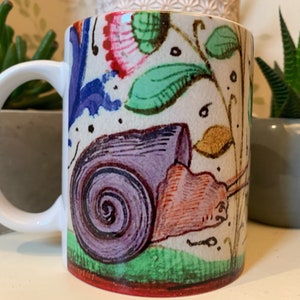 May include: White ceramic mug with a colourful illustration of a snail, featuring a purple shell and reddish-brown body. The design includes floral and botanical elements in blue, green, pink, and gold. The mug has a white handle and a red base.