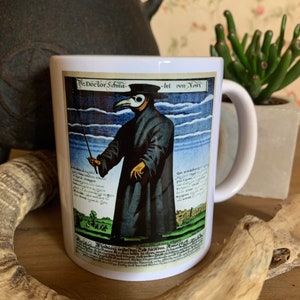May include: White ceramic mug with a black handle. The mug features a vintage illustration of a plague doctor wearing a long black coat, a beak mask, and a hat. The illustration is surrounded by text in German.