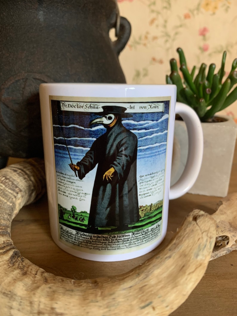 May include: White ceramic mug with a black handle. The mug features a vintage illustration of a plague doctor wearing a long black coat, a beak mask, and a hat. The illustration is surrounded by text in German.