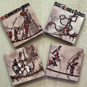 May include: Four square coasters with medieval illustrations. Each coaster depicts a different scene with rabbits in armour, wielding weapons, and interacting with other creatures. The coasters have a parchment-like background with text.