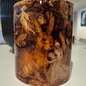 May include: A cylindrical ceramic vase with a detailed illustration of a brown hare, fairies, and forest foliage. The artwork is in warm, earthy tones, creating a whimsical and vintage aesthetic. The vase has a glossy finish.