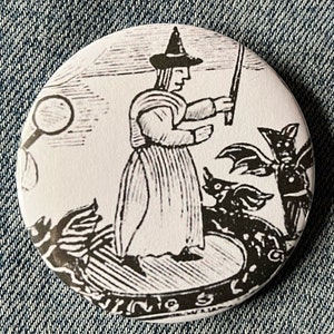 May include: Black and white illustration of a witch with a pointed hat and a wand, surrounded by bats and other creatures. The image is printed on a round button.