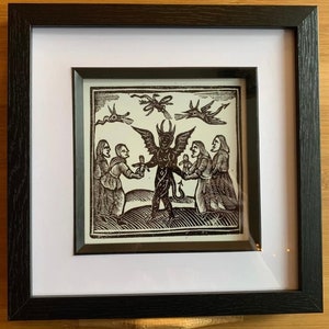 May include: A black and white woodcut print depicting a devil with wings and a tail, standing in the centre of the image. The devil is holding a small figure in its left hand and a cross in its right hand. Four people are standing around the devil, two on each side. The people are dressed in simple clothing and appear to be offering the devil the small figure. The print is framed in a black frame with a white mount.