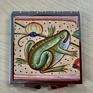 May include: A square, silver-framed compact mirror with a detailed illustration of a green frog. The frog is depicted in a medieval style, with a beige background and decorative floral elements.