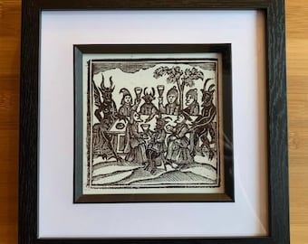 May include: A black and white woodcut print depicting a group of people and a devil seated around a table, drinking from goblets. The print is framed in a black frame with a white mat.