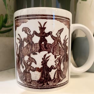 May include: White ceramic mug featuring a brown illustration of a circle of figures with horns and tails. The figures are dancing and holding hands. The mug has a handle on the right side.