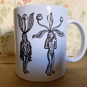 May include: White ceramic mug featuring a black and white illustration of two figures with plant-like heads. One figure is female, the other male. The mug has a curved handle and is set against a floral background.