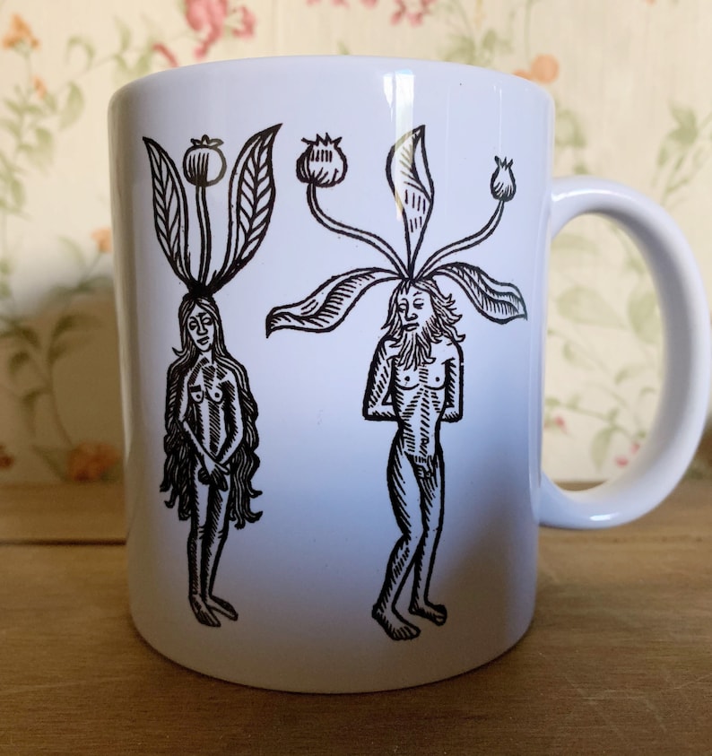 May include: White ceramic mug featuring a black and white illustration of two figures with plant-like heads. One figure is female, the other male. The mug has a curved handle and is set against a floral background.