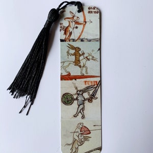 May include: A bookmark with a black tassel features four illustrations of rabbits in a medieval style. The top illustration shows a rabbit holding a bow and arrow. The second illustration shows a rabbit riding a horse. The third illustration shows a rabbit holding a sword and shield. The bottom illustration shows a rabbit holding a spear.