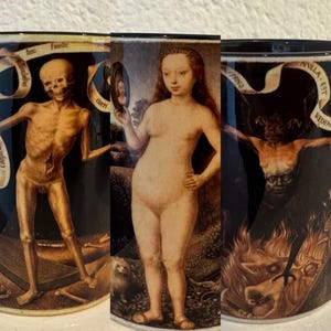 May include: Three black ceramic mugs with distinctive artwork. One mug shows a skeletal figure, another a nude woman holding a mirror, and the third depicts a demonic figure. Each mug has a dark background and detailed illustrations.