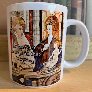 May include: White ceramic mug featuring a colourful image of a religious scene. The image depicts three figures, a man, a woman, and a child. The text "Let me just mansplain that to you..." is printed on the mug.