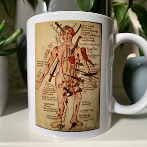 May include: A white ceramic mug featuring a vintage medical illustration of a human figure with multiple wounds and the text "Kamite aute forri remafiti atid Giare limni bed forme Camerofa Gardine ayanule fanti Tabsa bleerofa If bene mamemil Apofte fab Grame".