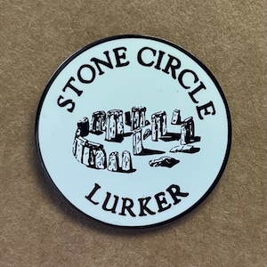 May include: A white enamel pin with a black outline. The pin features a black and white illustration of Stonehenge with the text "Stone Circle Lurker" around the image.