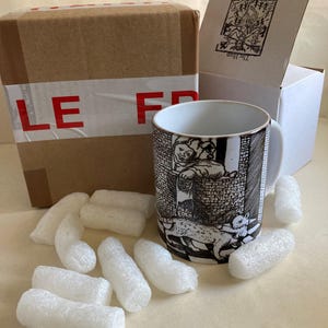 May include: A ceramic mug with a black and white illustration of a scene with a person and a cat. The mug is next to a brown cardboard box with red tape and a white box with text. White packing peanuts surround the mug.