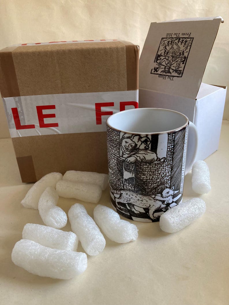 May include: A ceramic mug with a black and white illustration of a scene with a person and a cat. The mug is next to a brown cardboard box with red tape and a white box with text. White packing peanuts surround the mug.
