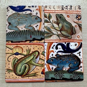 May include: A set of four square ceramic coasters, each with a medieval-style illustration of a frog. The frogs are in shades of green and blue, with detailed artwork and decorative borders. Each coaster measures approximately 10 cm by 10 cm.