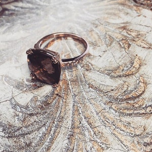 May include: A rose gold ring with a large, square-shaped, brown gemstone. The ring has a band of small diamonds.