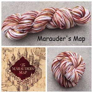 May include: A skein of yarn featuring a variegated color palette of white, deep red, and golden brown. The text Marauder's Map is displayed above the yarn. A graphic illustration of a castle with the text The MARAUDER'S MAP is also visible.