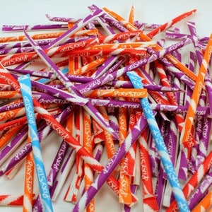 Può includere: Un grande mucchio di cannucce di caramelle Pixy Stix in vari colori tra cui viola, arancione, rosso e azzurro. Ogni sottile cannuccia di carta cilindrica presenta un motivo a righe diagonali e il marchio Pixy Stix stampato chiaramente sull'involucro.