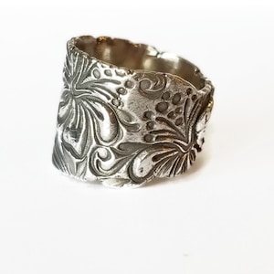 May include: A silver ring with a wide band featuring an intricate floral design. The band is textured and has a slightly irregular shape.