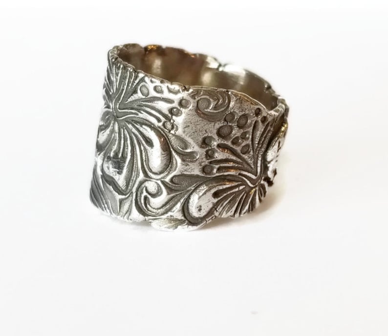 May include: A silver ring with a wide band featuring an intricate floral design. The band is textured and has a slightly irregular shape.