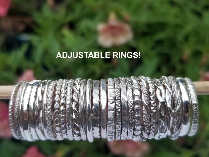 Pu&ograve; includere: Una collezione di anelli regolabili argentati con varie trame e motivi, esposti su un tassello di legno su uno sfondo verde e rosa sfocato. Il testo "ADJUSTABLE RINGS!" &egrave; visibile in alto.