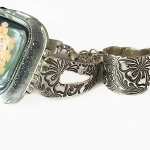 May include: A silver watch band and ring with a floral design. The watch band has a cutout in the center and the ring is wide.