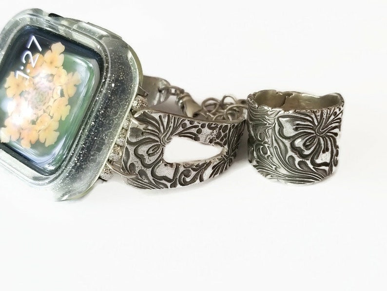 May include: A silver watch band and ring with a floral design. The watch band has a cutout in the center and the ring is wide.