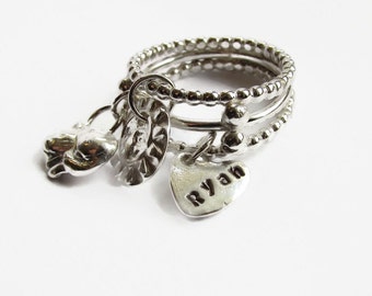 May include: Silver ring set with three bands and charms. The charms include a heart with the name "Ryan" engraved on it, a small hand, and a round charm with a textured design.