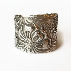 May include: A silver ring with a floral design. The ring is wide and has a textured surface.