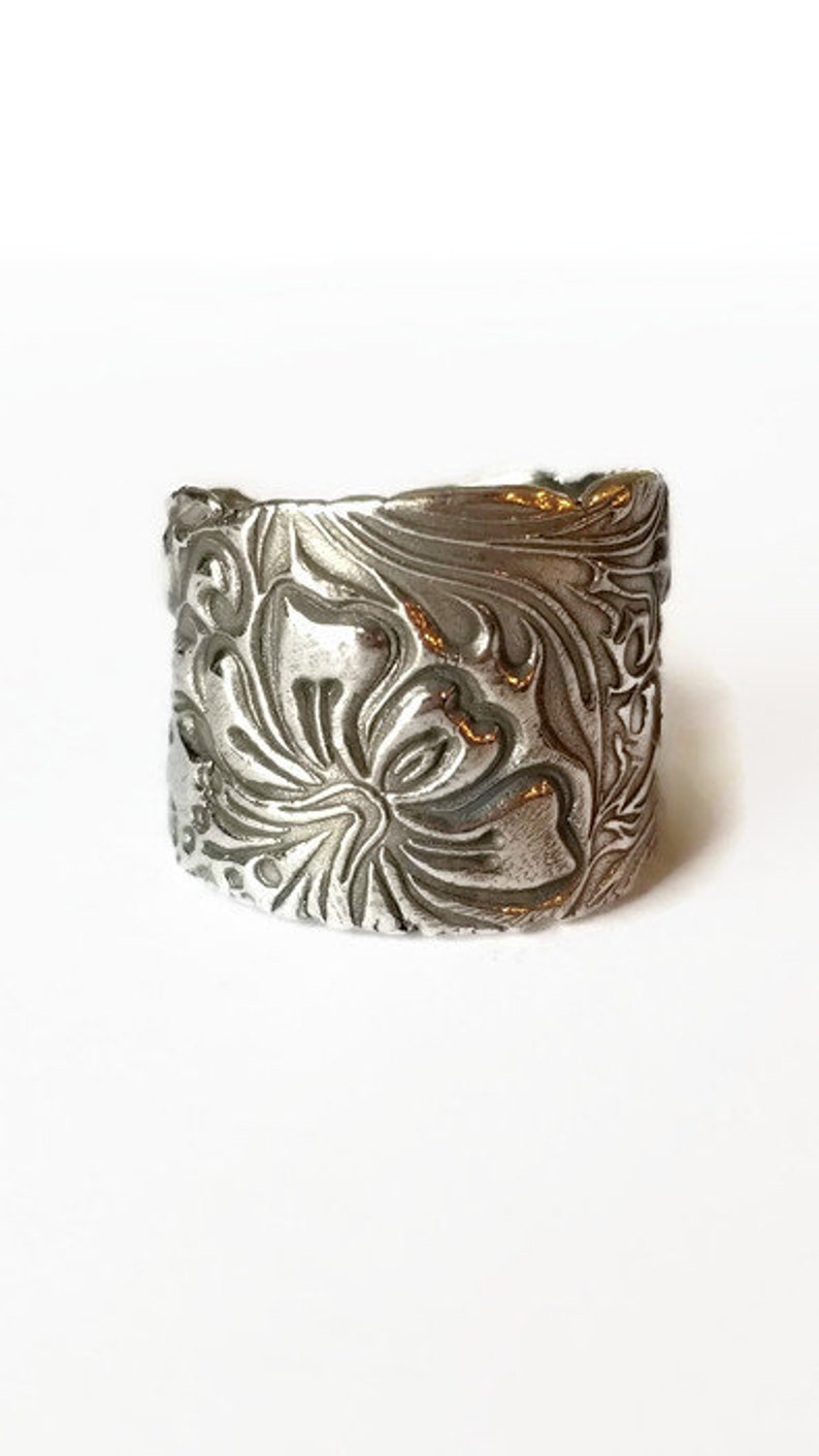 May include: A silver ring with a floral design. The ring is wide and has a textured surface.