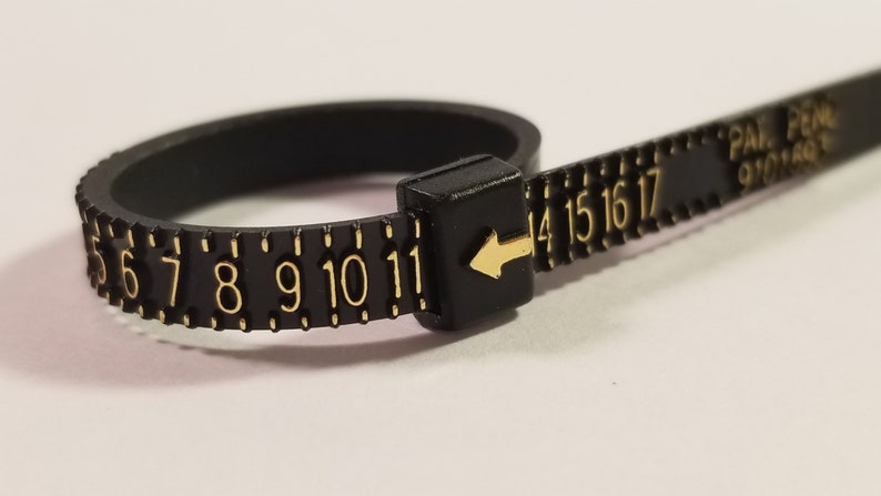 May include: Black plastic ring sizer with gold numbers from 5 to 17. The sizer has a black arrow pointing to the number 11.