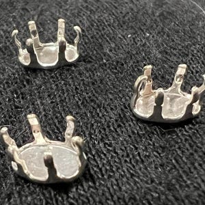 May include: Three silver-colored jewelry settings with a pronged design. The settings are round with multiple prongs extending upwards, designed to hold gemstones. The items are displayed on a dark, textured surface.