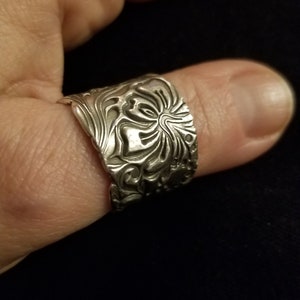 May include: A silver ring with a detailed floral design. The ring is wide and has a textured surface.
