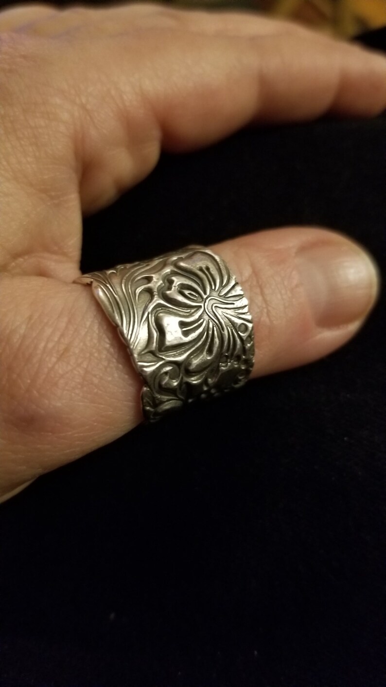 May include: A silver ring with a detailed floral design. The ring is wide and has a textured surface.