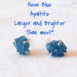 Peut inclure: Une paire de boucles d'oreilles avec des pierres d'apatite bleu néon serties dans de l'argent. Les pierres sont plus grandes et plus brillantes que la plupart. "Apatite bleu néon Plus grande et plus brillante que la plupart !" est écrit en bleu au-dessus des boucles d'oreilles.