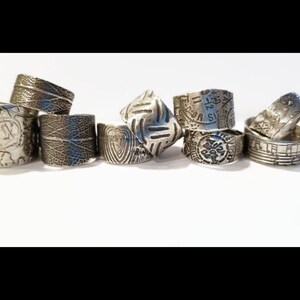 May include: A collection of eight silver rings with different textured designs. The rings are stacked and arranged on a white surface.