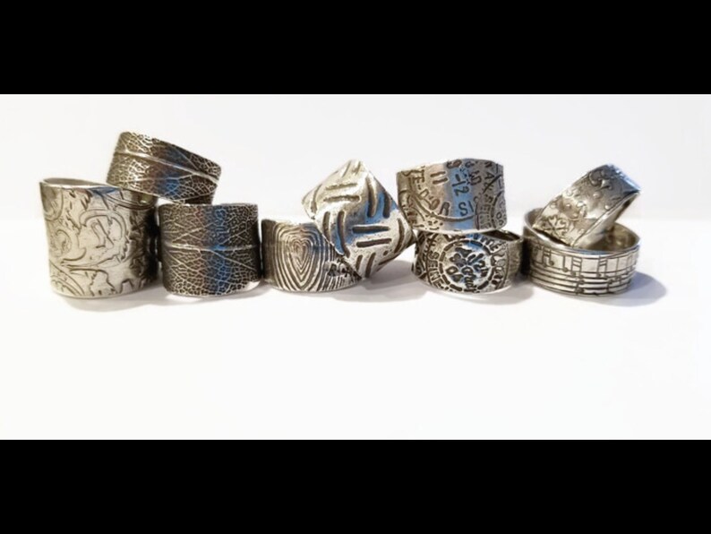 May include: A collection of eight silver rings with different textured designs. The rings are stacked and arranged on a white surface.