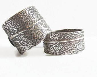 May include: Two silver-toned rings with a leaf vein pattern. One ring is wide and wraps around, while the other is a band. The rings have a textured surface and are set against a white background.