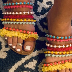 Pu&ograve; includere: Sandali in pelle marrone con pi&ugrave; cinturini colorati con perline e nappe. I cinturini presentano accenti turchesi, rosa, bianchi, rossi, gialli e arancioni. I sandali hanno un anello per le dita e sono indossati su un tappeto fantasia.