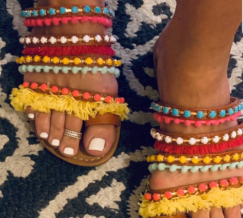 Pu&ograve; includere: Sandali in pelle marrone con pi&ugrave; cinturini colorati con perline e nappe. I cinturini presentano accenti turchesi, rosa, bianchi, rossi, gialli e arancioni. I sandali hanno un anello per le dita e sono indossati su un tappeto fantasia.