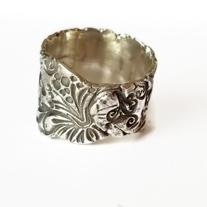 May include: A silver ring with a wide band featuring an intricate, embossed floral design. The band is textured and has a slightly irregular shape.