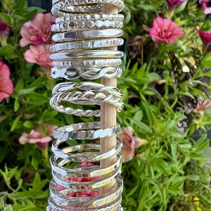 Pu&ograve; includere: Una collezione di braccialetti d'argento esposti su un tassello di legno. I braccialetti presentano vari design, tra cui motivi intrecciati, attorcigliati e strutturati. Lo sfondo presenta fiori rosa e fogliame verde, creando un ambiente naturale e vibrante.