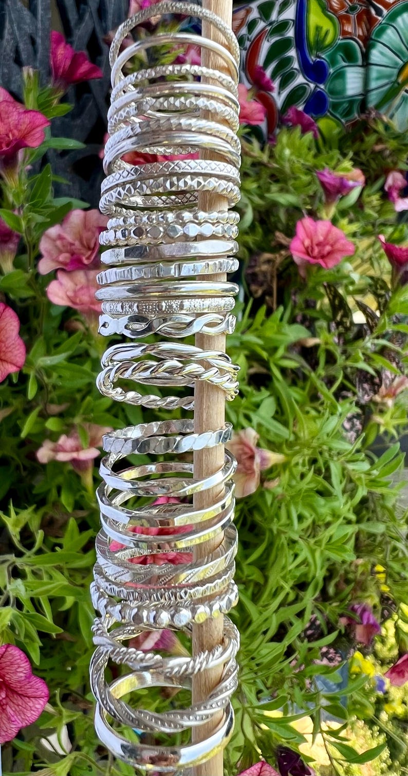 Pu&ograve; includere: Una collezione di braccialetti d'argento esposti su un tassello di legno. I braccialetti presentano vari design, tra cui motivi intrecciati, attorcigliati e strutturati. Lo sfondo presenta fiori rosa e fogliame verde, creando un ambiente naturale e vibrante.