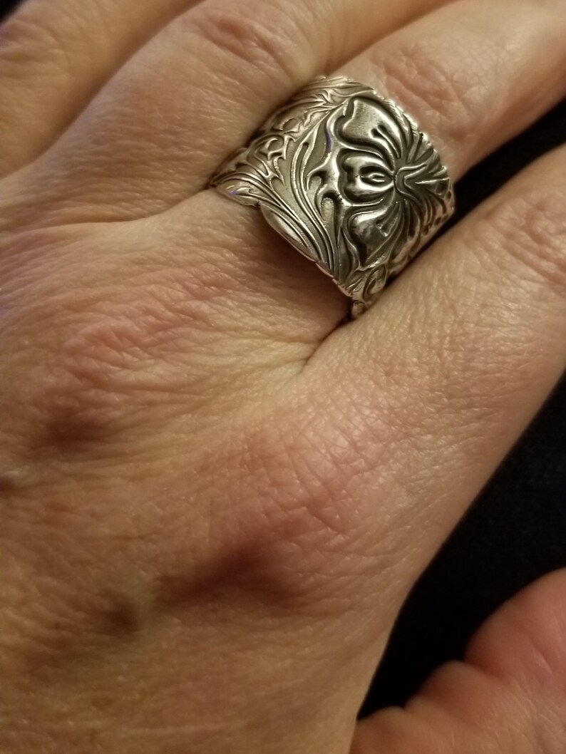 May include: A silver ring with a floral design. The ring is wide and has a detailed pattern.