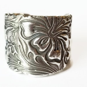 May include: A silver ring with a floral design. The ring is wide and has a textured surface.