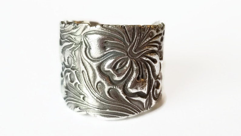 May include: A silver ring with a floral design. The ring is wide and has a textured surface.