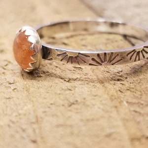 Puede incluir: Un anillo de plata con un diseño de rayos de sol y una piedra preciosa redonda de color naranja.
