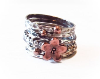 May include: A silver ring with multiple bands, each with a hammered texture. The ring is adorned with small copper beads and a copper flower charm with the letter "R" engraved on it.